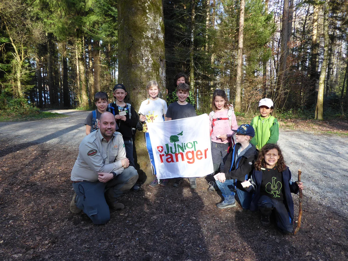 Mit dem Junior-Ranger-Programm lernen Kinder und Jugendliche, sorgfältig mit der Umwelt umzugehen. Eine Gruppe von Kindern posiert im Wald mit der Fahne des Junior-Ranger-Programms.